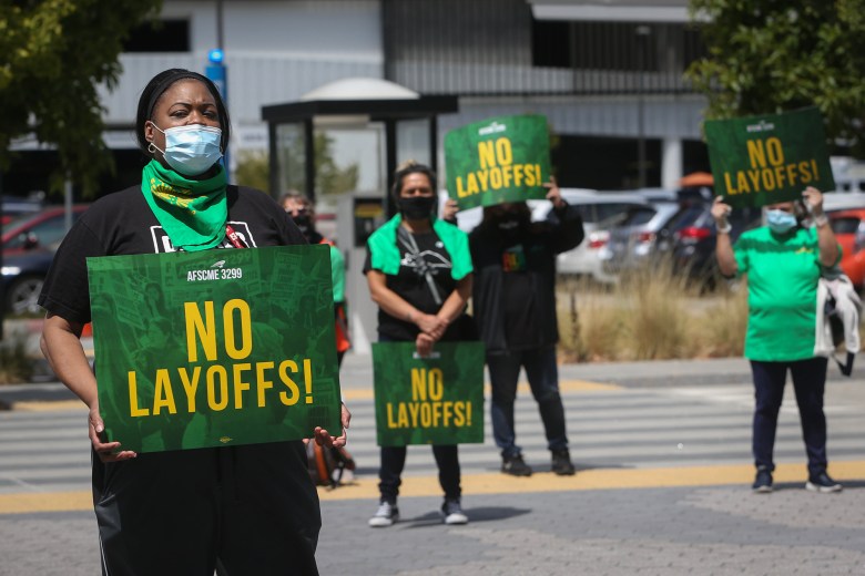 Protesters standing outdoors holding green signs with the words, "NO LAYOFFS!" in yellow and "AFSCME 3299." The setting is a protest.