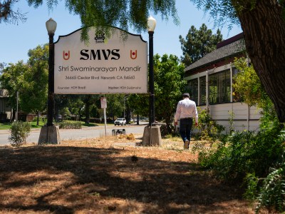 A person wearing a white button-up shirt walks past a sign for a hindu temple located on the front lawn of a building.