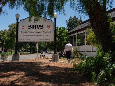 A man walks away looking down as he walks past a sign that reads, "SMVS Shri Swaminarayan Mandir" and a building.