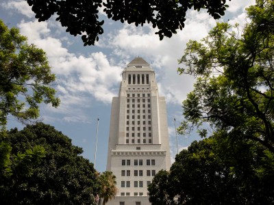 LA City Hall is framed by lush green trees and a partly cloudy sky, standing tall in the center with a clear view of its tiered architecture. The foliage in the foreground adds depth and contrast to the modern lines of the building.