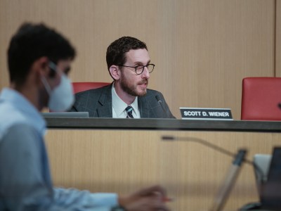 A person wearing glasses, sits behind a dais in a grey suit. A name tag next to to them read "Scott D. Wiener." In the left corner, a person is out of focus while wearing a face mask.