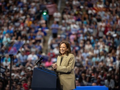 Vice President Kamala Harris, wearing a tan blazer and black shirt, stands behind a podium with a view of a crowded arena behind her at a campaign rally.