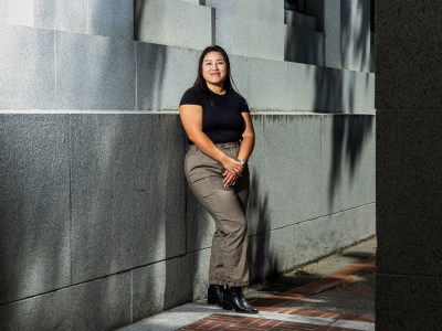 A person in grey slacks, black boots and a black shirt stands in the corridor between two buildings. Dramatic light causes and interesting contrast of light and shadows.