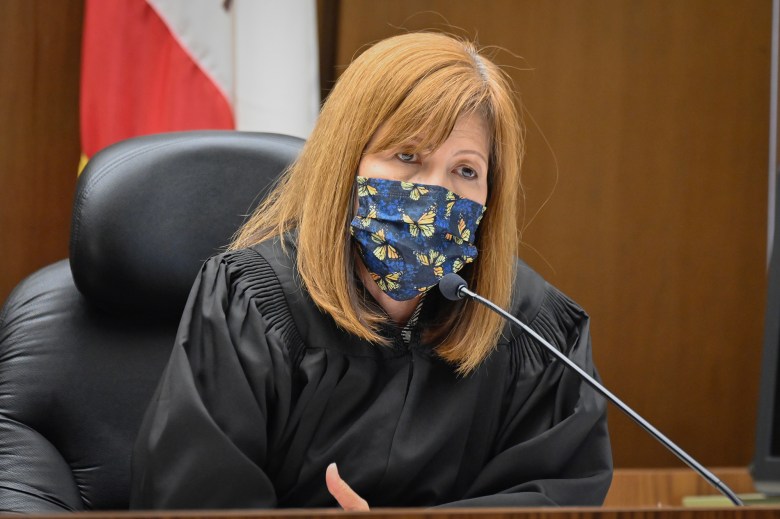 A person seated at a judicial bench, wearing a black robe and a patterned face mask with butterfly designs. A microphone is positioned in front of them, and the background includes a partially visible flag and wood-paneled walls, indicating a courtroom setting. The individual appears to be speaking or listening attentively.