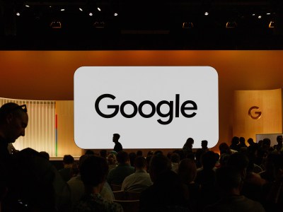 Silhouettes of people seated in a large audience, facing a stage with a prominent screen displaying the Google logo in bold black text on a white background. The stage is lit with warm lighting, creating a soft glow, while the room is dimly lit around the audience.