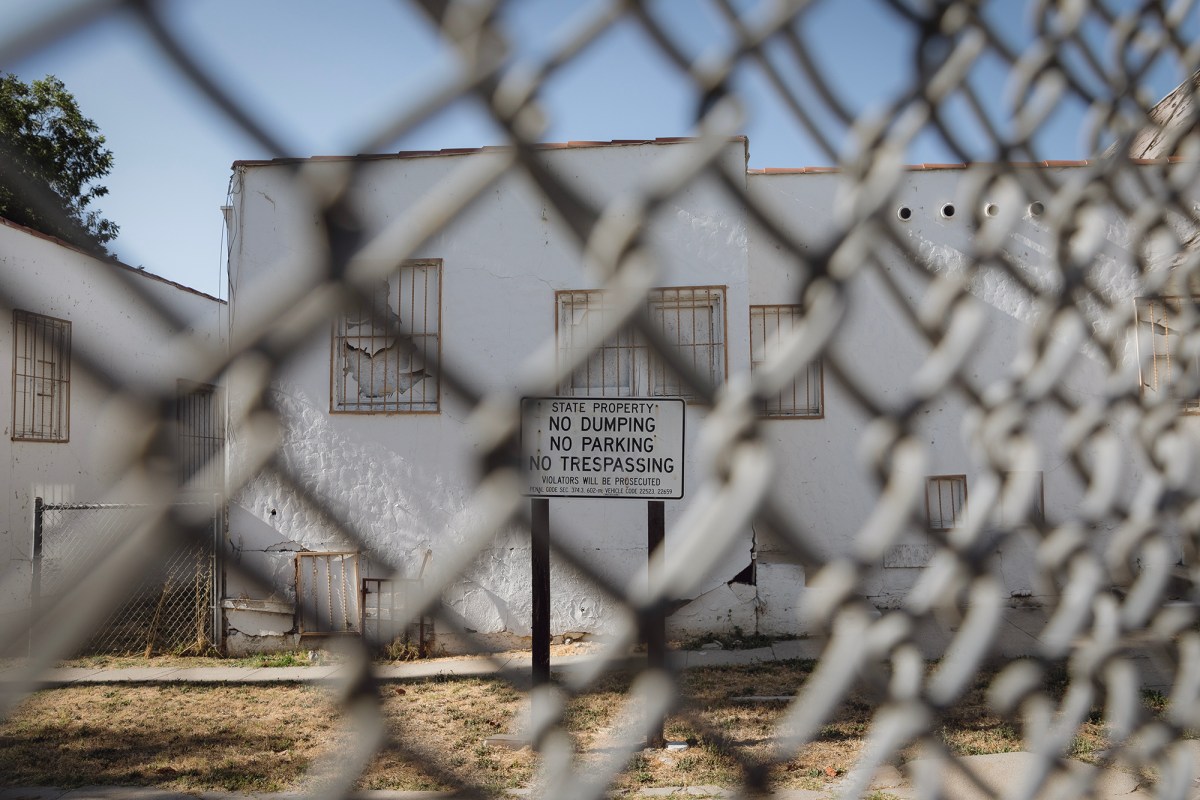 A "No Dumping. No Parking. No Trespassing." sign sits behind a double-chain-linked fence. A dilapidated, white, one-story building sits behind the sign. The area looks abandoned.