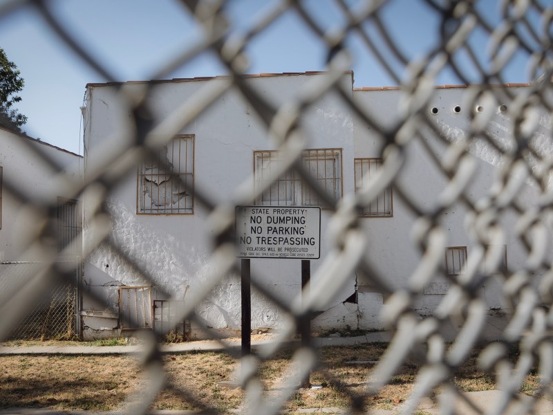 A "No Dumping. No Parking. No Trespassing." sign sits behind a double-chain-linked fence. A dilapidated, white, one-story building sits behind the sign. The area looks abandoned.