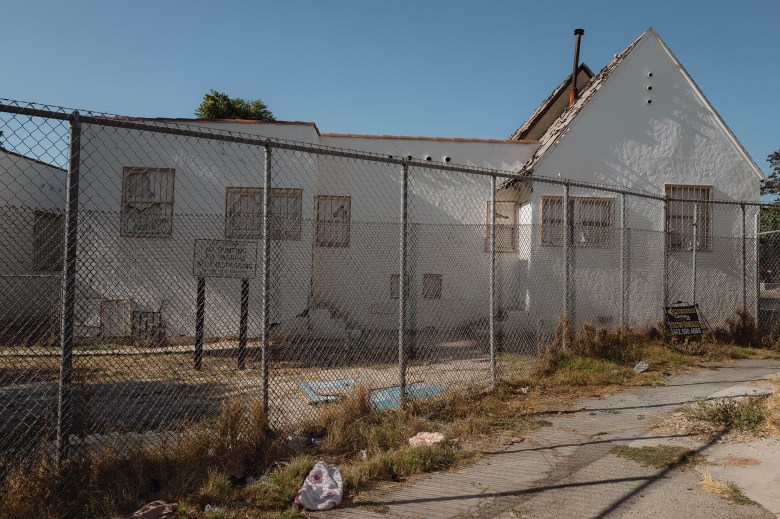 A "No Dumping. No Parking. No Trespassing." sign sits behind a double-chain-linked fence. A dilapidated, white, one-story building sits behind the sign. The area looks abandoned.