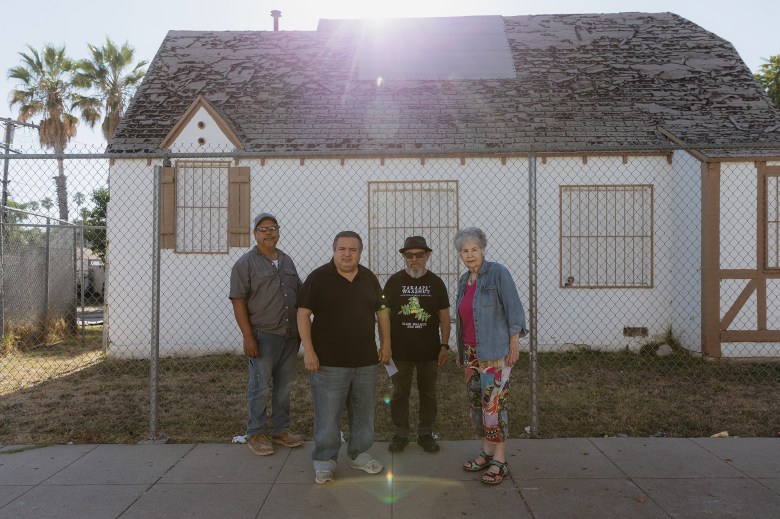 Saul Contrera, Raymond Gutierrez, Roberto Flores and Joanne Nickolas stand in front of a chain-linked fence that surrounds a property where an abandoned, single-story, white building sits.