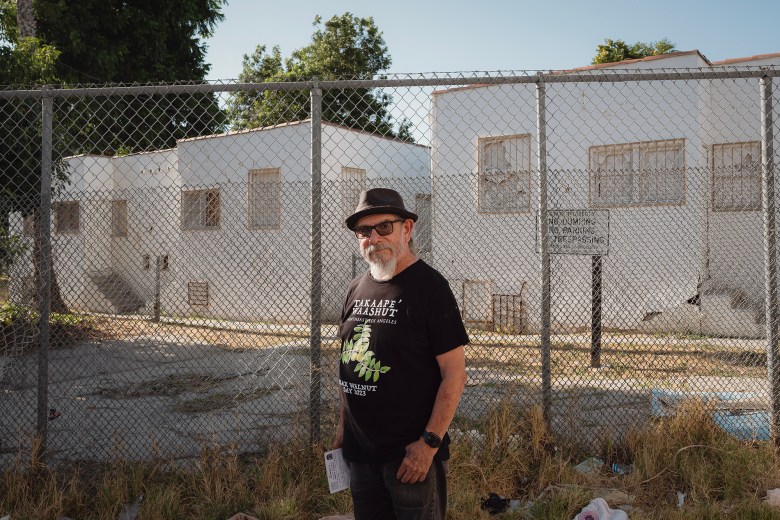 Roberto Flores, wearing a black hat, t-shirt and pants, stands in front of an abandoned building. The building is sits behind two chain-linked fences. A "No Trespassing" sign sits on the property, behind the fence.