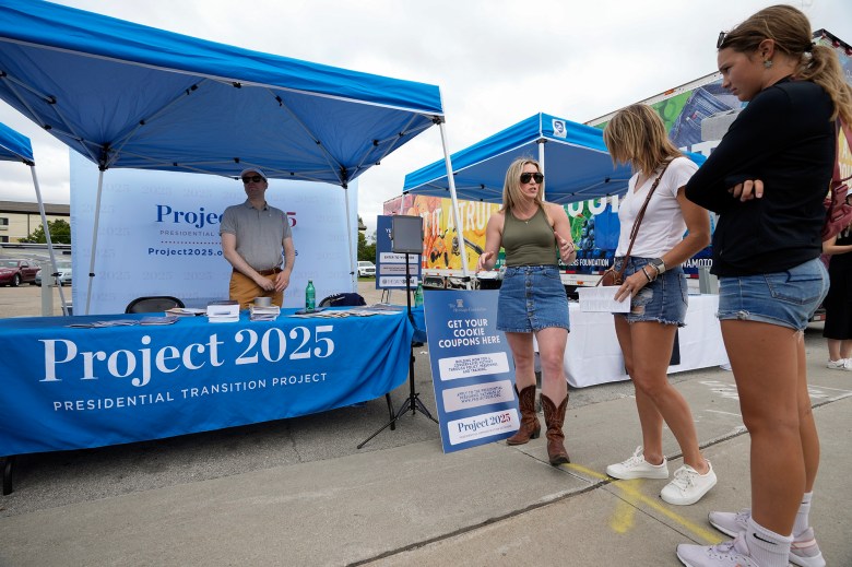 A person in a green tank top and denim skirt talks to two people in denim shorts at the Project 2025 booth, which has blue tents and a table covered with a Project 2025 banner. Another person stands behind the table with brochures, while a sign in front reads, "Get your cookie coupons here."