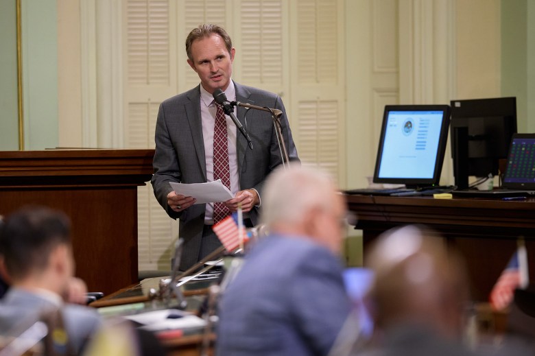 A lawmaker wearing a grey suit and red tie stands in front of a microphone stand while holding a piece of paper as they speak to other lawmakers.