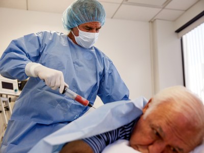 A doctor, wearing blue scrubs a bouffant cap, and a face mask, uses a syringe on a patient's lower back as they lay down on a medical bed at a clinic.