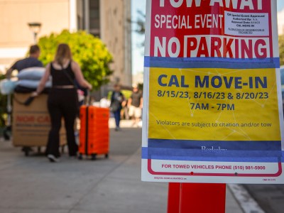 "A close-up view of a sign taped to a pole that reads 'CAL MOVE-IN' with dates 8/15/23, 8/16/23, and 8/20/23 from 7 AM to 7 PM, indicating no parking during these times due to move-in events. The background shows a blurry scene of people, including a person pushing a cart with large boxes and an orange suitcase, as they navigate a busy sidewalk.