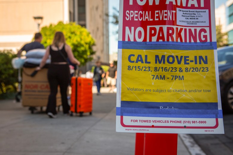 "A close-up view of a sign taped to a pole that reads 'CAL MOVE-IN' with dates 8/15/23, 8/16/23, and 8/20/23 from 7 AM to 7 PM, indicating no parking during these times due to move-in events. The background shows a blurry scene of people, including a person pushing a cart with large boxes and an orange suitcase, as they navigate a busy sidewalk.