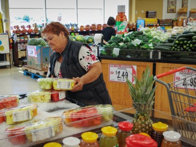 An older individual stands in a grocery store near a display of pre-packaged fruit containers on ice, selecting a pack of watermelon and other mixed fruits. The person is wearing a floral shirt under a dark vest. Behind them are avocados on sale, with a price sign for $2.49 per avocado. A shopping cart with a pineapple inside is visible nearby, along with other fresh produce such as cucumbers.