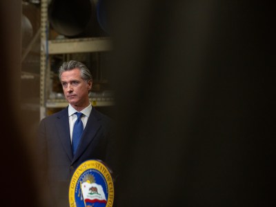 Viewed through a narrow, black sliver, Gov. Gavin Newsom stands at a podium while at a press conference.