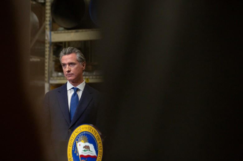 Viewed through a narrow, black sliver, Gov. Gavin Newsom stands at a podium while at a press conference.