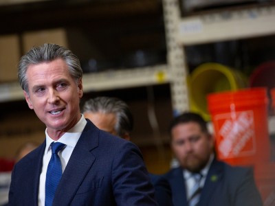 A tilted view of Gov. Gavin Newsom wearing a navy blue suit and looking to his right side while other lawmakers stand behind him at a Home Depot location.