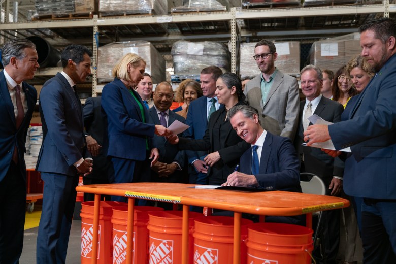 Surrounded by smiling state lawmakers and other stakeholders, Gov. Gavin Newsom signs legislation while seated at a "desk" made of a large industrial roller cart at a Home Depot retail store.