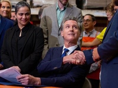 Flanked by state lawmakers, Gov. Gavin Newsom shakes another lawmakers hand in celebration while seated at a desk at a Home Depot in San Jose.