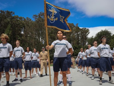 A group of individuals standing in formation outdoors on a bright, sunny day. The person at the front of the group holds a blue flag with a gold anchor and rope emblem, displaying a confident posture. The group wears matching gray T-shirts and navy blue athletic shorts, with trees and a blue sky in the background.