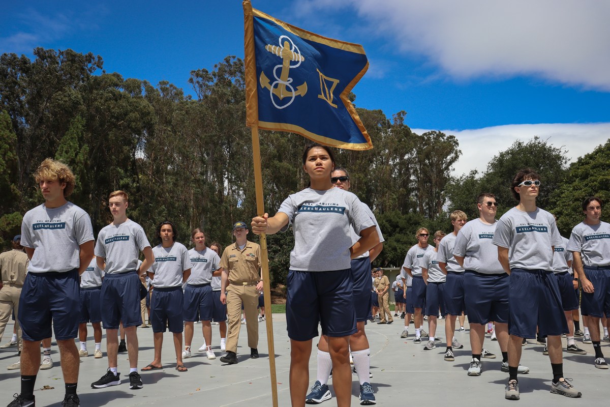 A group of individuals standing in formation outdoors on a bright, sunny day. The person at the front of the group holds a blue flag with a gold anchor and rope emblem, displaying a confident posture. The group wears matching gray T-shirts and navy blue athletic shorts, with trees and a blue sky in the background.