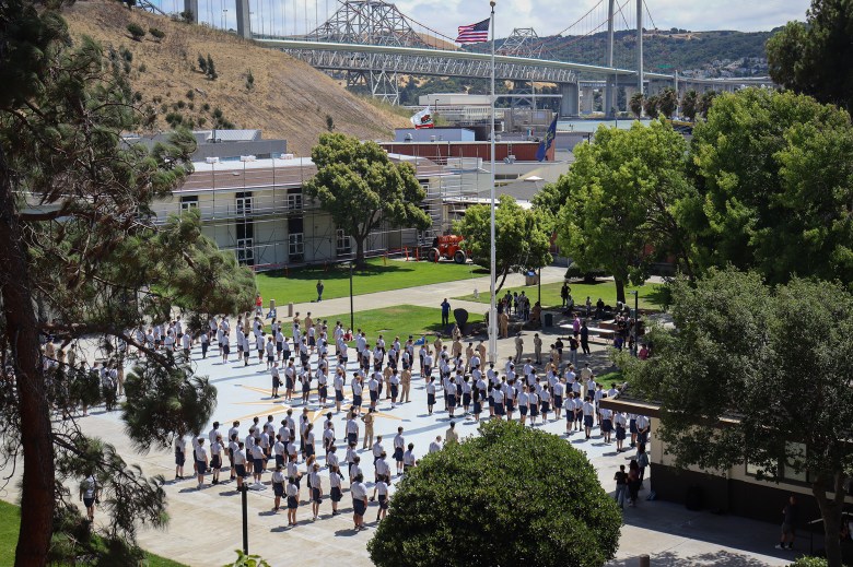 A wide view of a campus assembly, with numerous individuals standing in uniform rows on a paved courtyard surrounded by green lawns and trees. Buildings with scaffolding are visible in the background, along with a tall flagpole displaying the American flag and a nearby bridge spanning across the horizon. The scene is bright and sunny, highlighting the organized outdoor gathering.