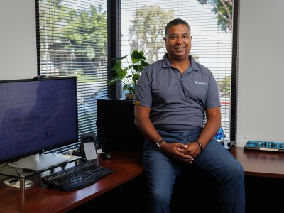 A person wearing a grey polo shirt and blue jeans, sits on a desk, while a computer monitor stands on the desk, in an office.