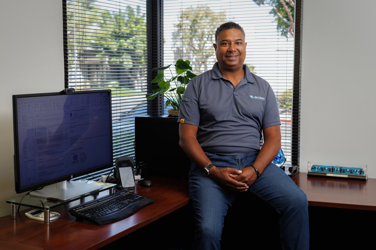 A person wearing a grey polo shirt and blue jeans, sits on a desk, while a computer monitor stands on the desk, in an office.