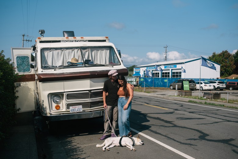 A man and woman embrace with their dog laying in front of them while they stand near an RV parked on the side of the road.