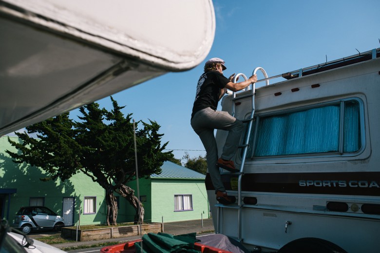 A man climbs the back ladder of an RV to get on top of the roof while its parked next to a street.