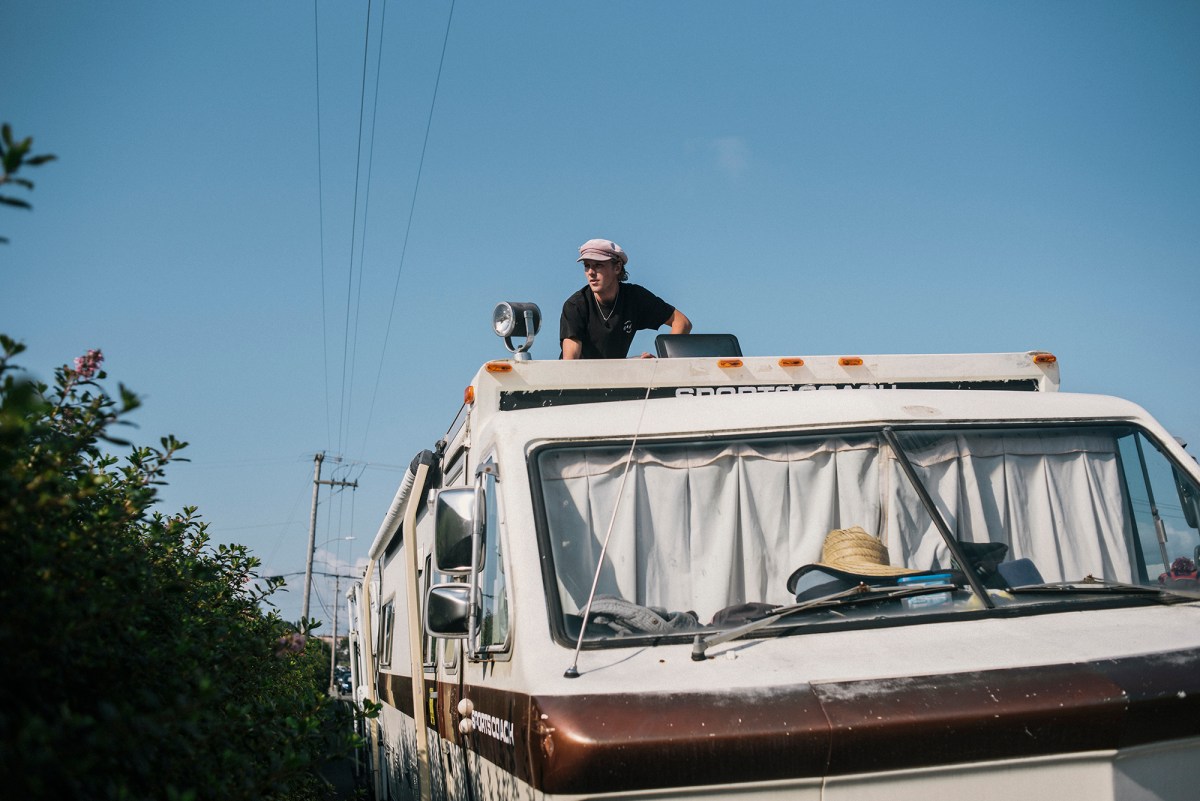 A man with a hat on top of an RV to check the roof while it's parked next to a street.