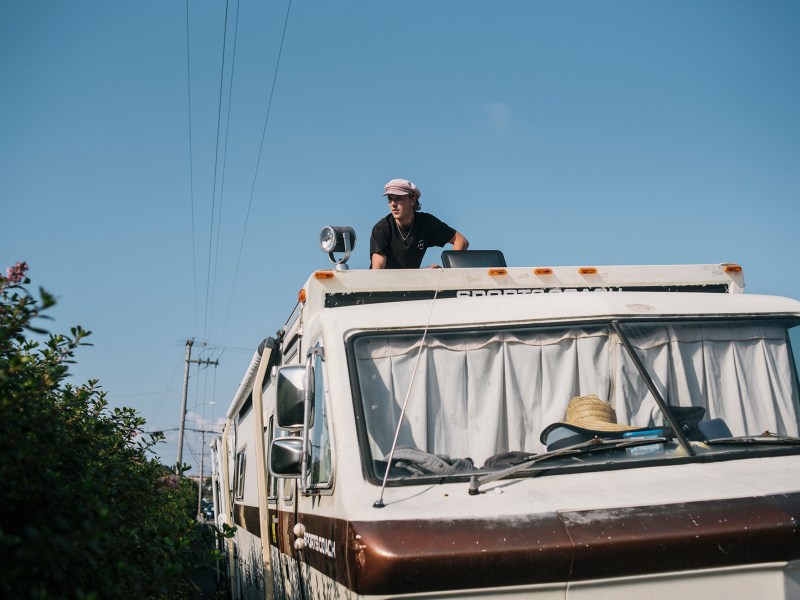 A man with a hat on top of an RV to check the roof while it's parked next to a street.