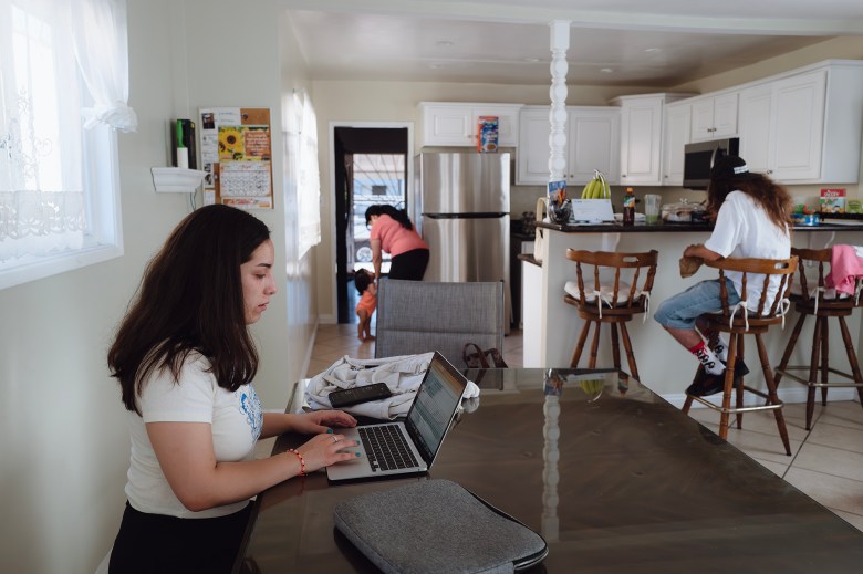 A person sits at a table using a laptop, focusing on the screen. Behind them, a kitchen scene unfolds: another individual with long hair and a cap sits on a high stool at the kitchen counter, eating. In the background, a third person tends to a small child near an open door. The room is bright, with white cabinets and a stainless steel refrigerator in the kitchen, and a window with lace curtains on the left.