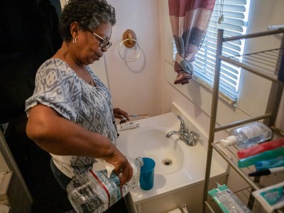 A woman standing in front of a faucet in a bathroom pours herself a cup of water of water from a container to brush her teeth without using contaminated water