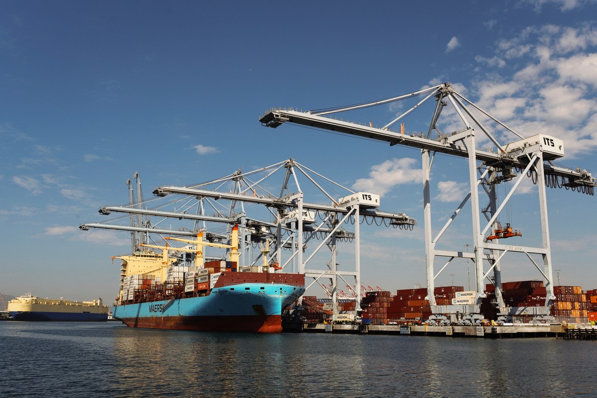 A blue and burnt red cargo ship is unloaded by various cranes at a port. Metal shipping containers are seen in the port. The ship is floating in the water.