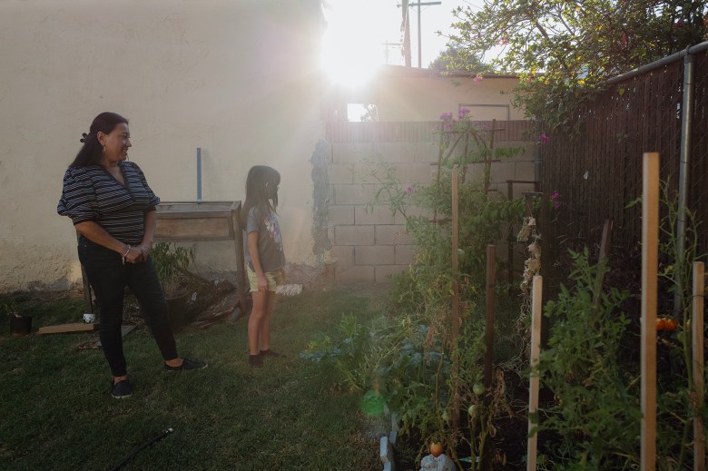 A person dressed in a striped black and white shirt stands in a garden with a child dressed in a grey t-shirt and yellow shorts. Sun rays peak through the edge of the roof of a building, creating sun flares in the frame. A vegetable garden is in front of the duo.