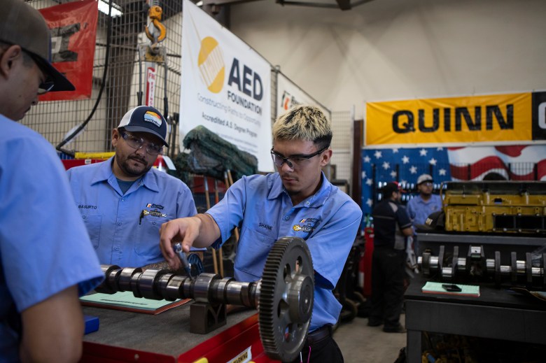 A student in a mechanic's outfit and safety goggles measures a part of a tractor engine on a table in a mechanic's classroom with other students watching.