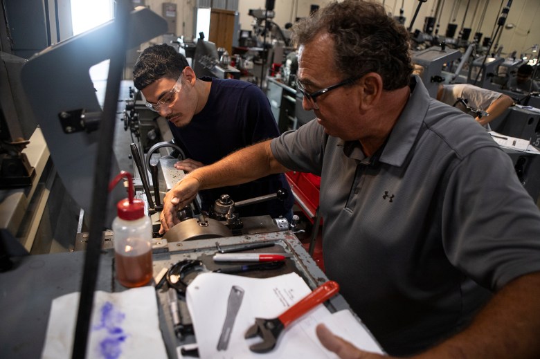 A teacher and student are wearing safety goggles and looking inside a machine to show how it works.