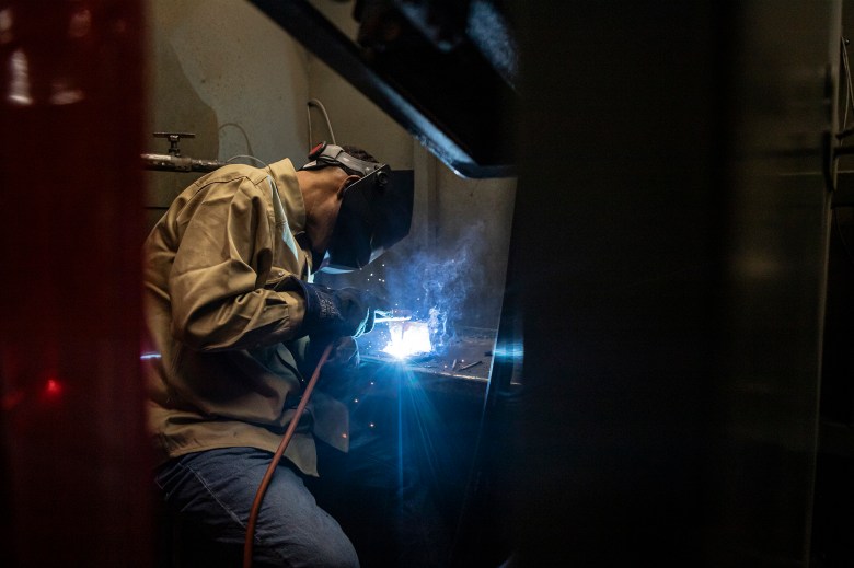 A student in welding mask and other safety gear welds a piece of metal in a work station in a college classroom.