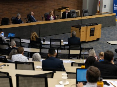 Four people, a mix of lawmakers and officials, sit behind a dais in a conference hall with attendees.