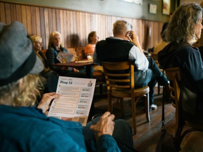 A person wearing a grey hat with a black band holds a paper with the words "Prop 33" printed on it. The scene is a bar, with other patron drinking beer and sitting at tables.