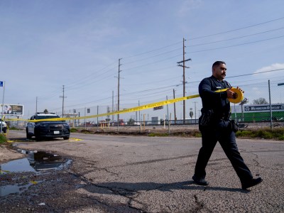 A police officer is unrolling a roll of crime scene tape along an empty road next to a fence and train track to create a crime scene.