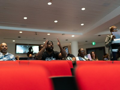 People sit in rows of red theater chairs during an event in Sacramento. One person in the center holds both hands up in frustration while speaking.