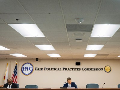 Two people sit spaced apart at a long, wooden table in a conference room with "Fair Political Practices Commission" displayed on the wall behind them. The room is lit by several ceiling lights, and the setup includes microphones and nameplates for each individual. The California state flag and the U.S. flag are positioned to the side, next to a sign for the FPPC. The individuals appear to be reviewing documents or taking notes. The overall atmosphere is formal and administrative.