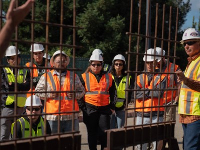 Students in yellow and orange safety vests and white safety helmets stand outside and watch two instructors as part of an iron workers training program.