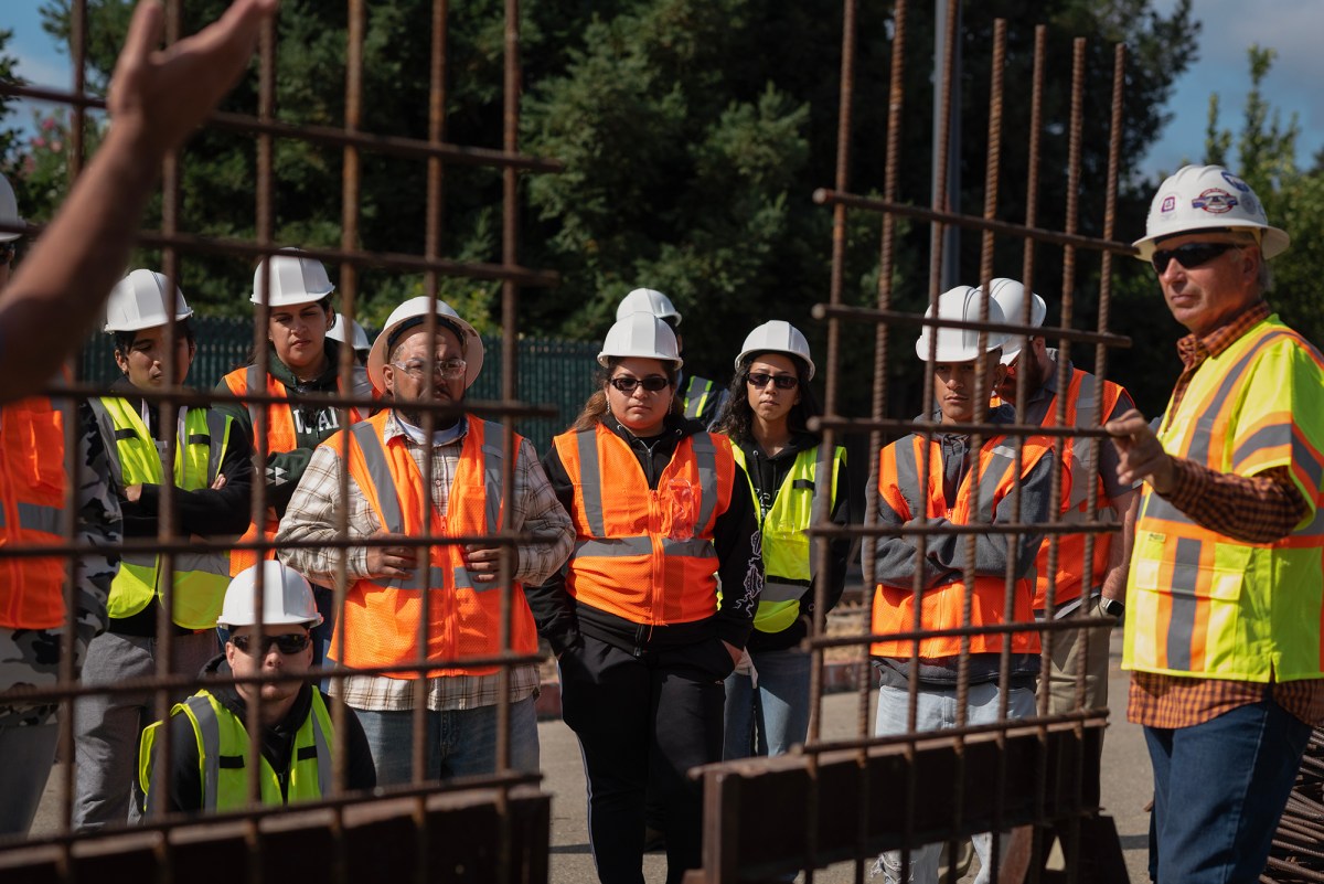 Students in yellow and orange safety vests and white safety helmets stand outside and watch two instructors as part of an iron workers training program.