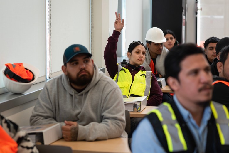 Students in yellow and orange safety vests sit at desks in a classroom. A young woman, wearing a yellow safety vest, raises her hand to ask a question.