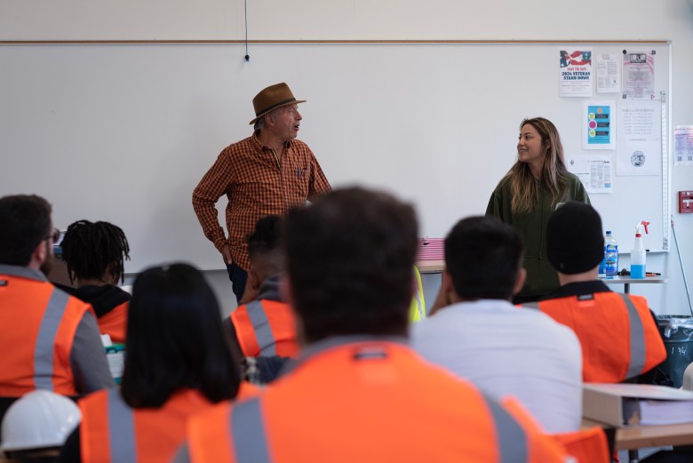 Students in orange reflective safety vests sit in a classroom as they listen to two teachers at the front of a class. The class is part of a Trades Orientation Program.